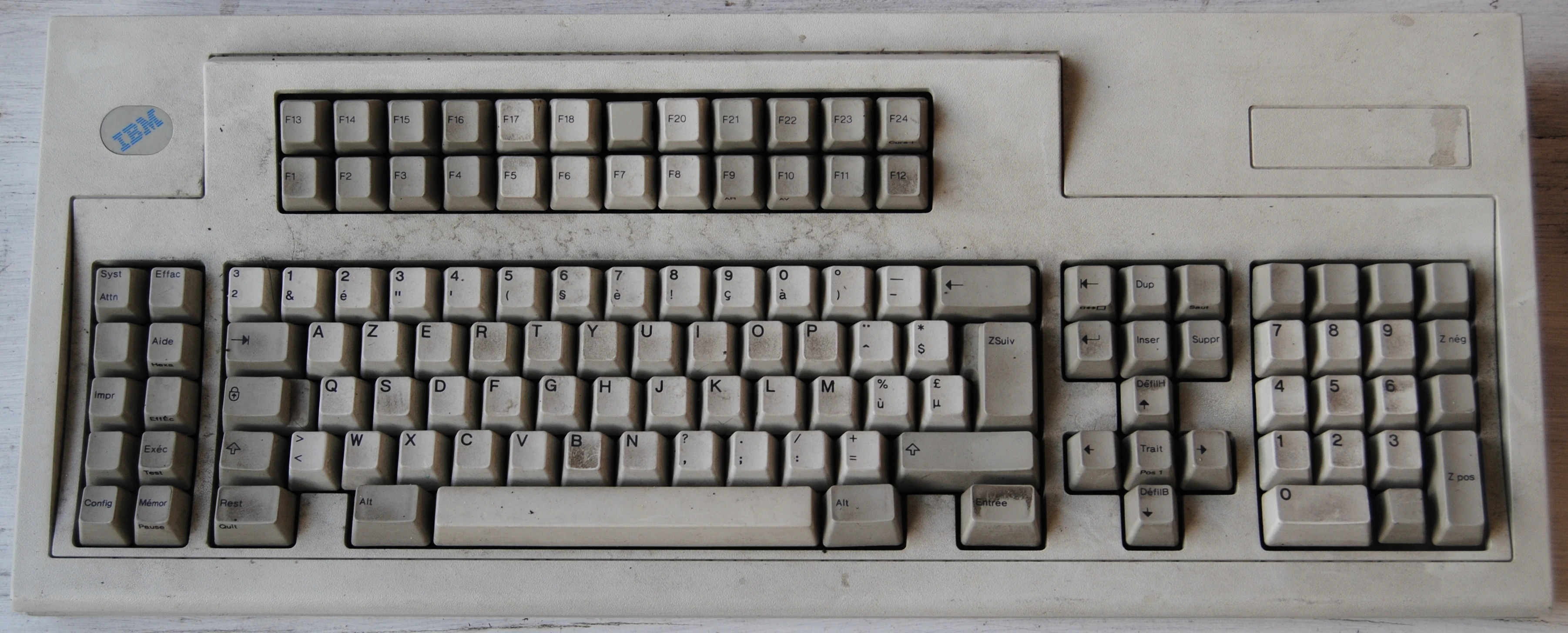 Photograph of the keyboard, with a dirty grayish plastic case. It's an old AZERTY French layout, without CTRL or Super keys. The Enter key is named "ZSuiv" (ZNext), and at the usual Right CTRL location there is a key labelled "Entrée" (Enter). Above the usual 12 function keys is another row up to F24. There is an arrow block with a central "Trait" key. There is a numpad. At the left is a 2-column block with strangely labelled special keys.